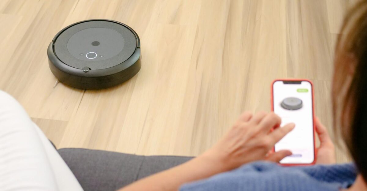 A person uses a smartphone app to control a robotic vacuum on a hardwood floor showcasing smart home technology