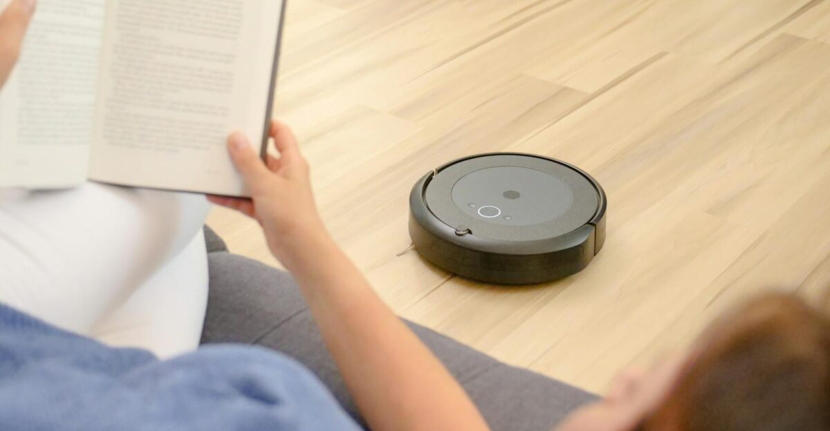 A woman relaxes on a sofa reading while a robotic vacuum navigates the floor