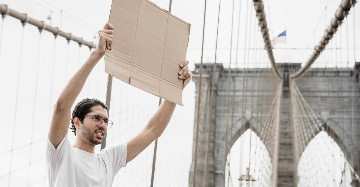 A man stands on Brooklyn Bridge holding a blank cardboard sign symbolizing protest and activism