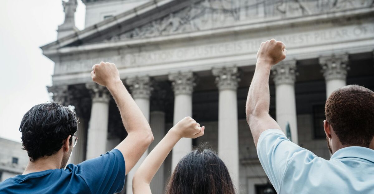 Three people raising fists in protest outside a government building with Greek-style architecture