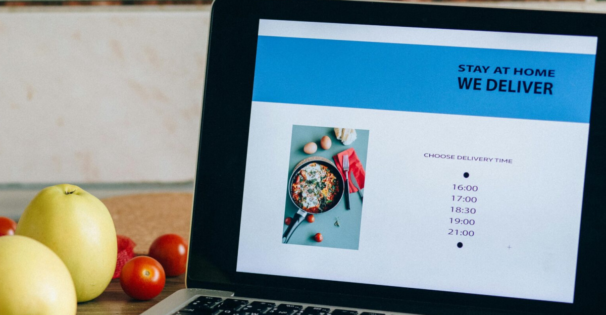 Laptop showing a delivery app beside fresh fruits and vegetables on a kitchen counter.