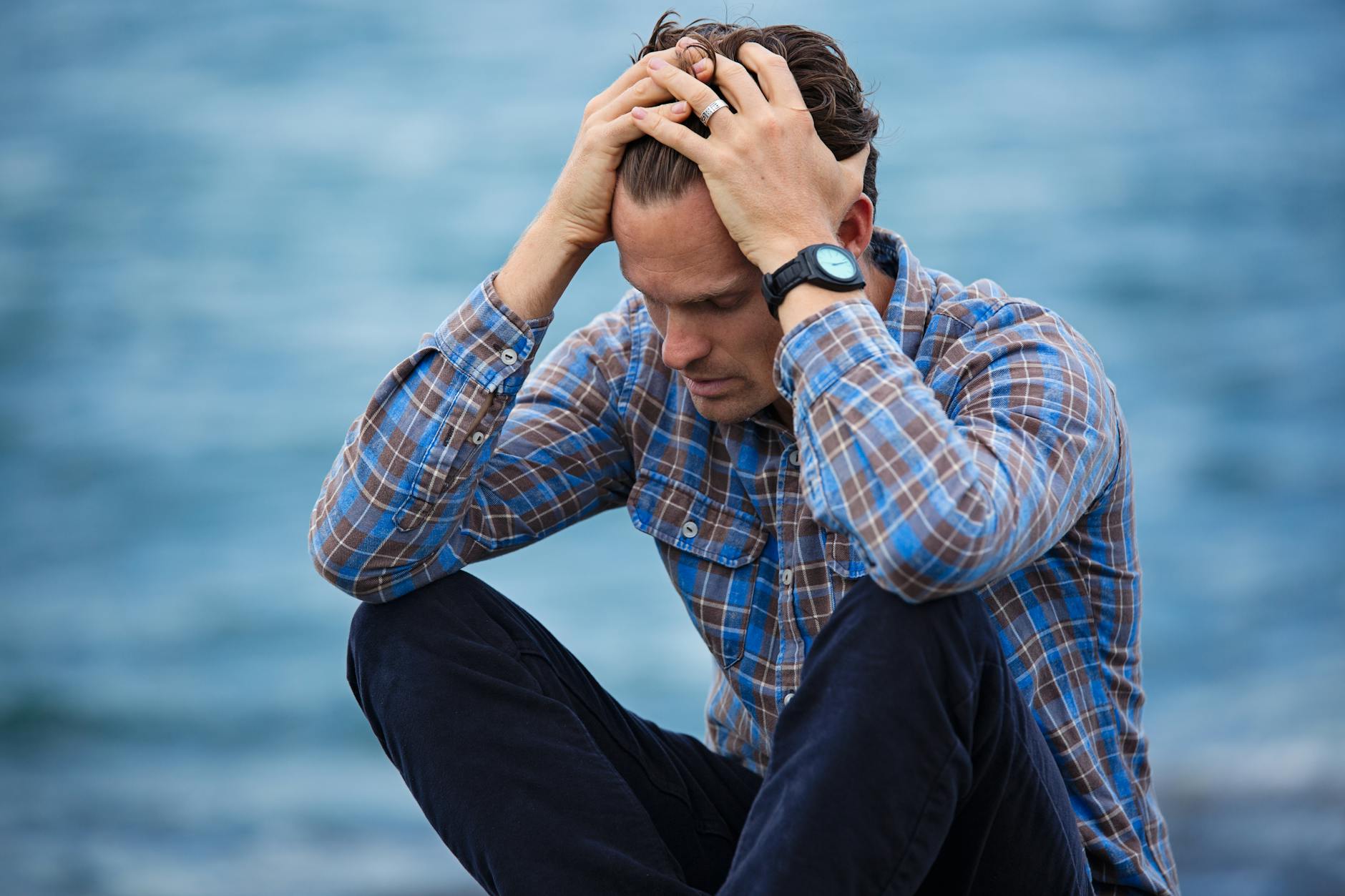 A man in a plaid shirt sits by the water looking distressed symbolizing stress