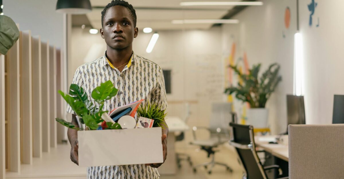 A serious man carries a box of belongings in an office setting symbolizing dismissal or job change