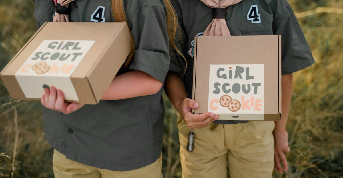 Two girl scouts holding cookie boxes with visible logos in an outdoor setting