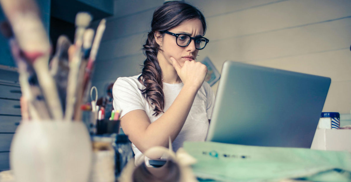 Young woman with glasses deeply focused on a laptop surrounded by art supplies in a home office.