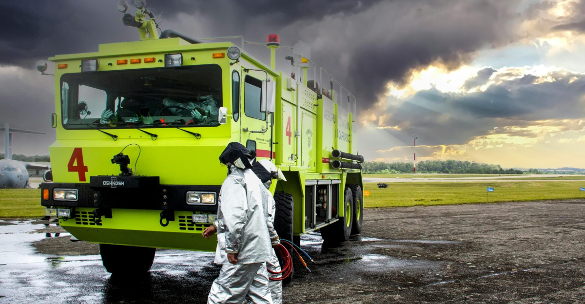 Firefighters in protective gear near a fire truck on a runway with dark storm clouds.