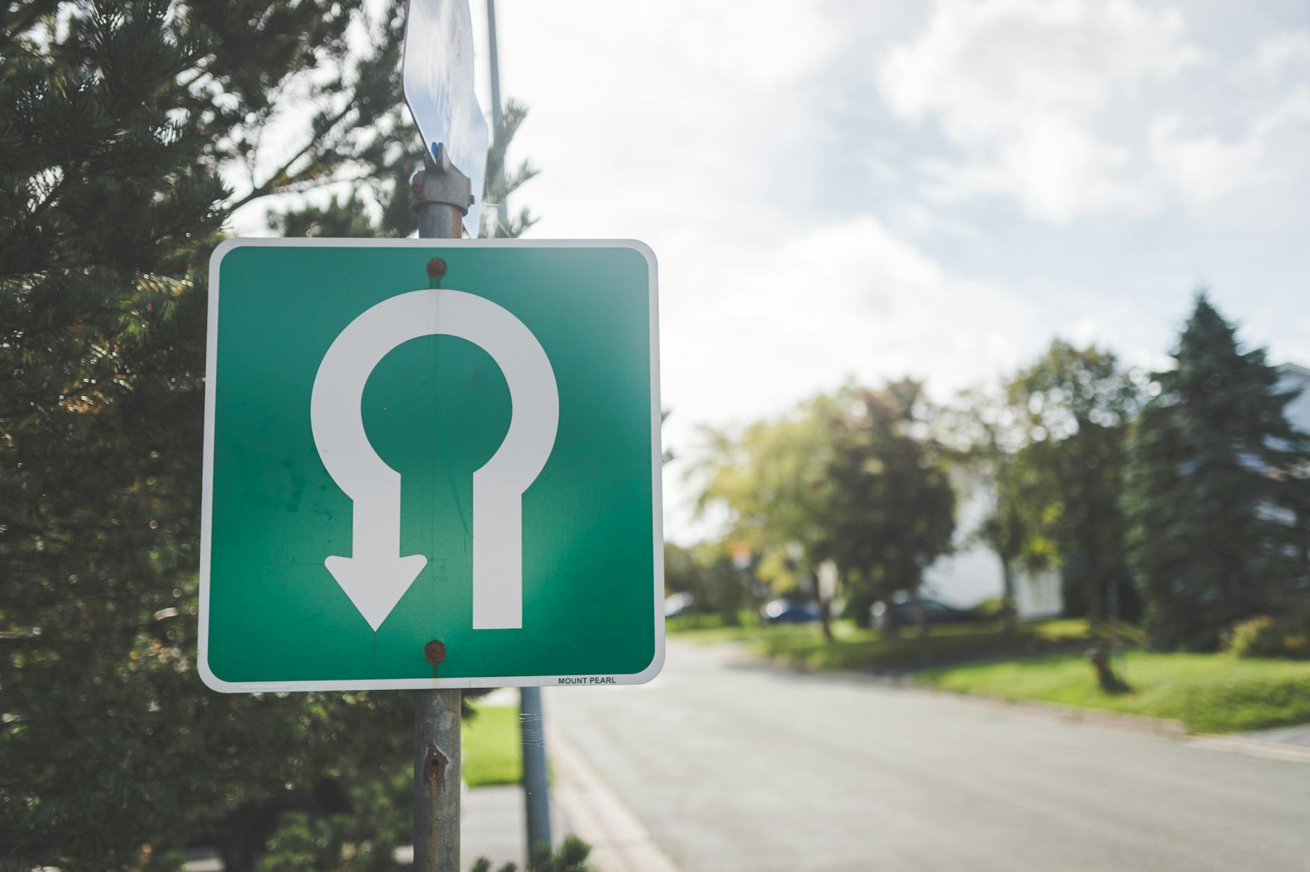 Close-up of a green U-turn road sign on a sunny suburban street with trees