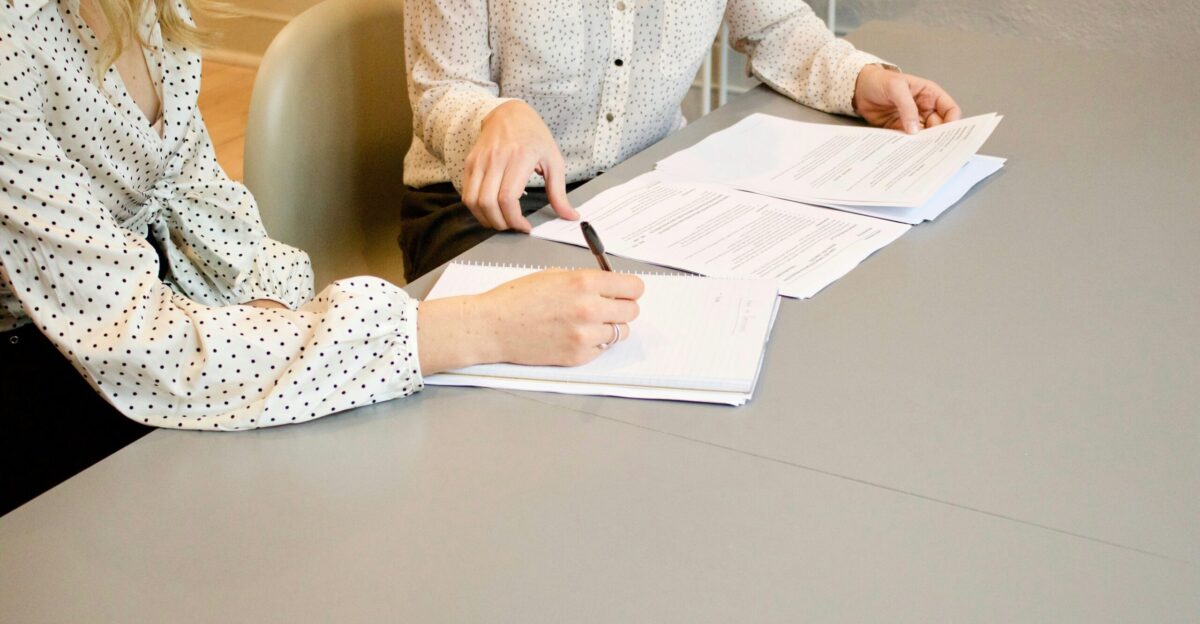 woman signing on white printer paper beside woman about to touch the documents