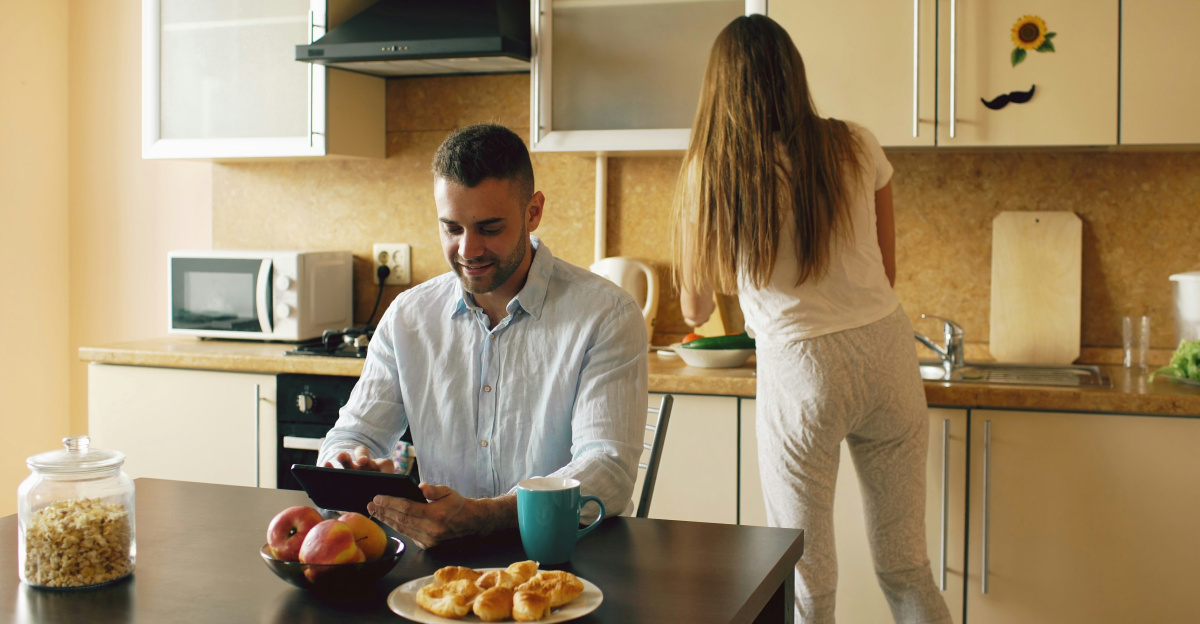 Couple in kitchen, man using tablet, woman preparing food.