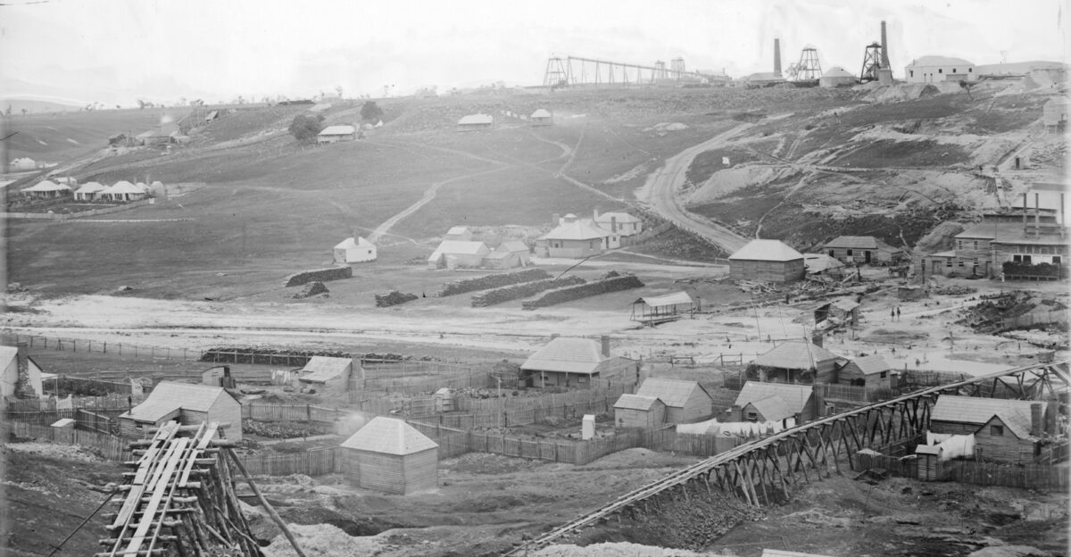 Overlooking township shows houses some with washing on the line gold mines some with towers chimney stacks wooden track l l supported by logs