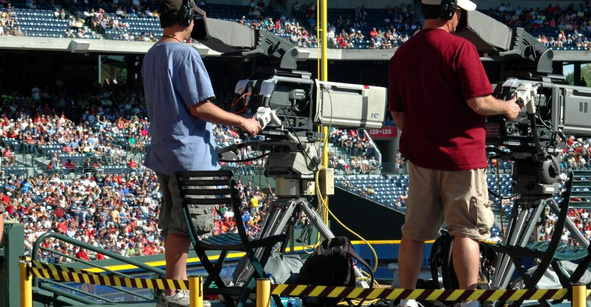 Camera operators capturing a live sports event at a crowded outdoor stadium filled with spectators