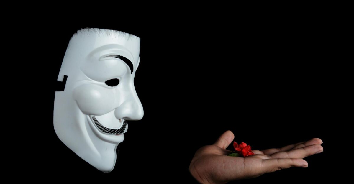 Side view of an anonymous mask facing a hand holding a red flower against a black background