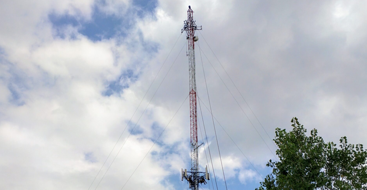 The image captures a guyed lattice mast, commonly referred to as a tower, outfitted with T-Mobile US mobile/cellular phone panel antennas supporting various telecommunication technologies including LTE, HSPA, UMTS, EDGE, and GSM (4G/3G/2G). The specific panel antennas installed on this site are the Ericsson AIR 21 and a CommScope antenna. During the time the photo was taken, the site was undergoing maintenance, with a tower technician/climber visible in the frame.