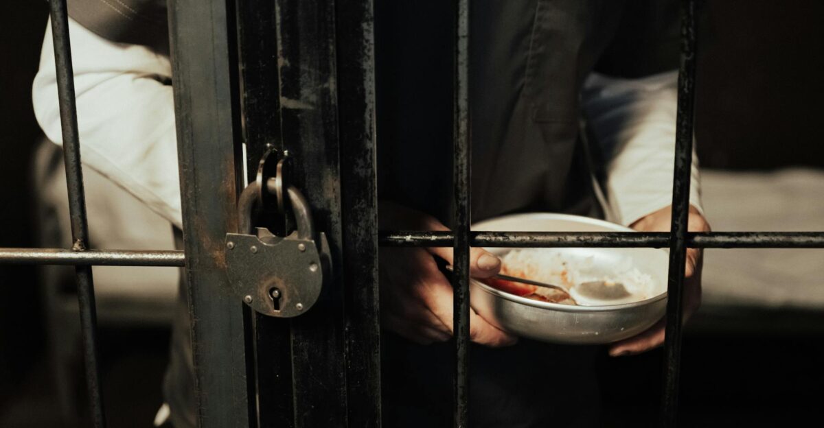 Close-up of a prisoner holding a bowl behind iron bars illustrating confinement