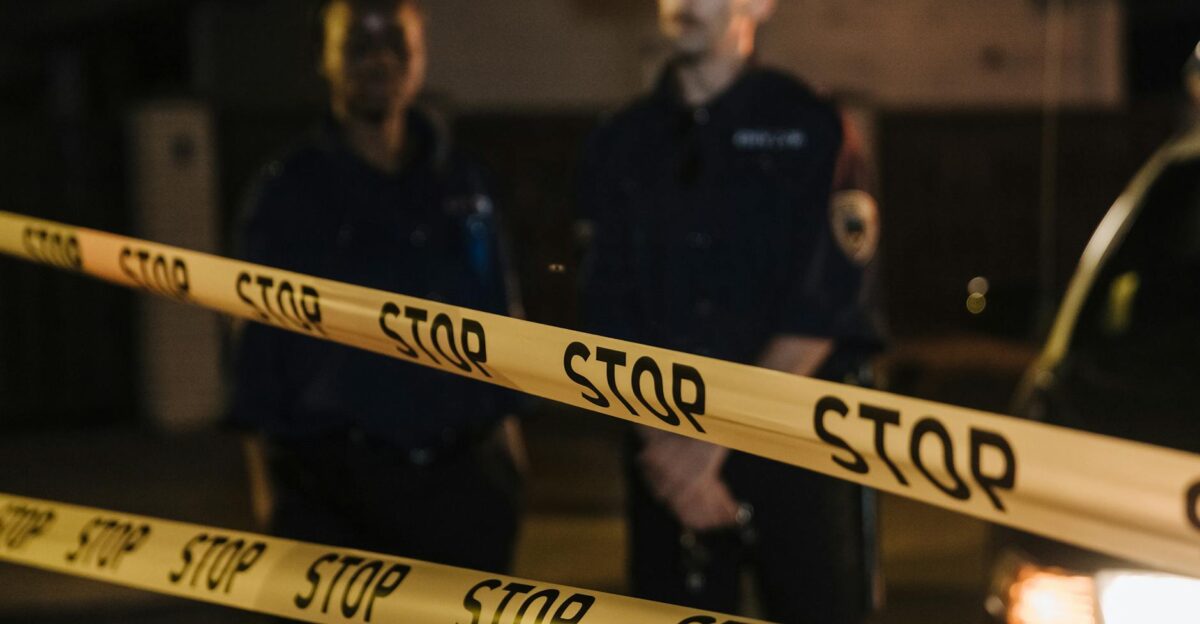 Two police officers standing behind a stop tape at night scene partially illuminated by car headlights