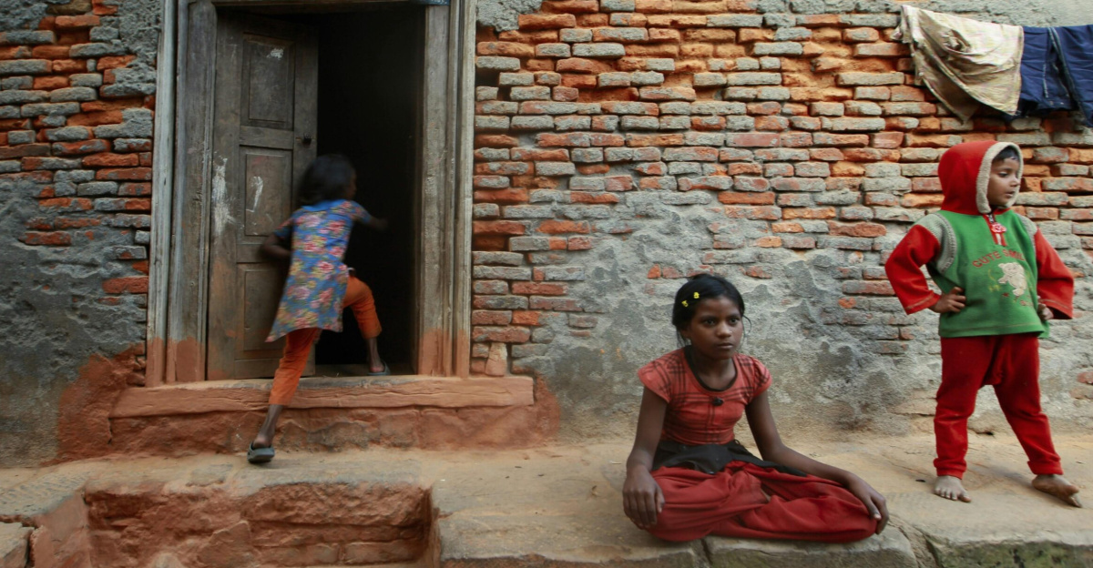 Children play around a rustic brick house with visible wear and poverty themes.