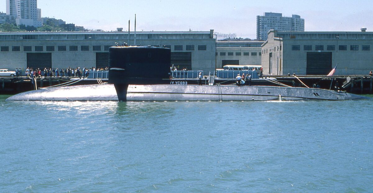 A naval submarine docked at an urban harbor with people on shore