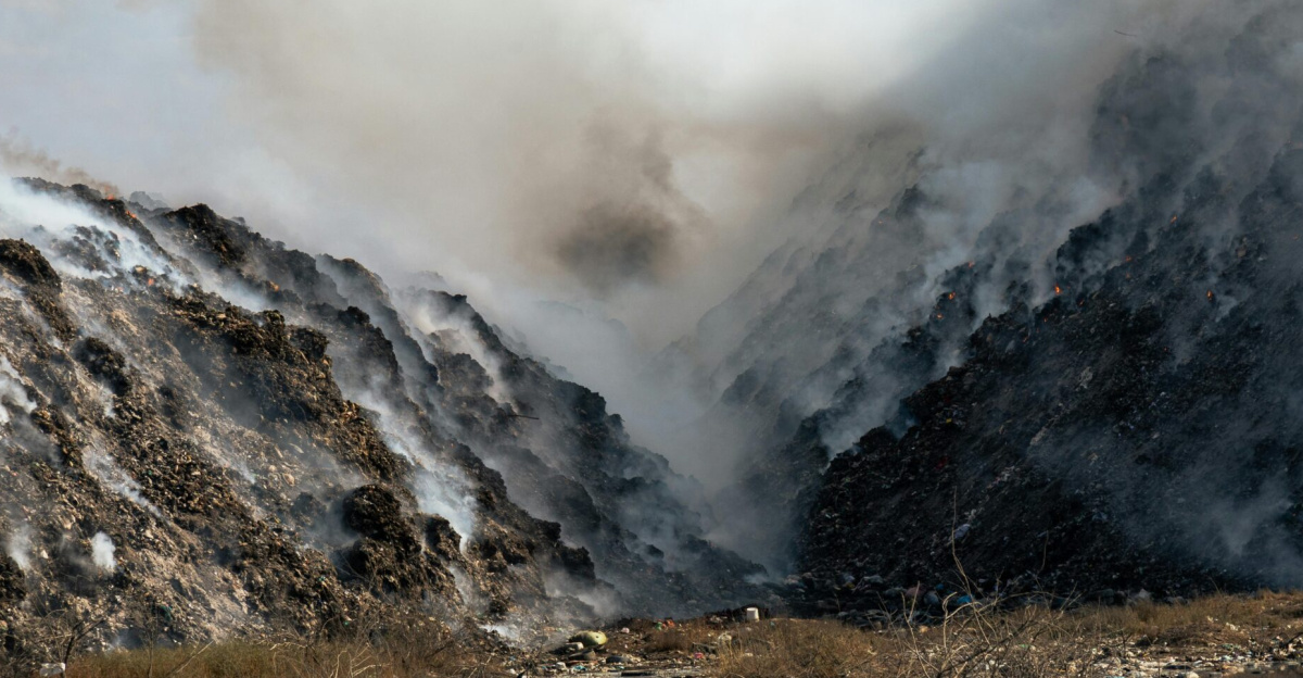 Thick smoke billows from a burning landfill surrounded by piles of waste and trash.
