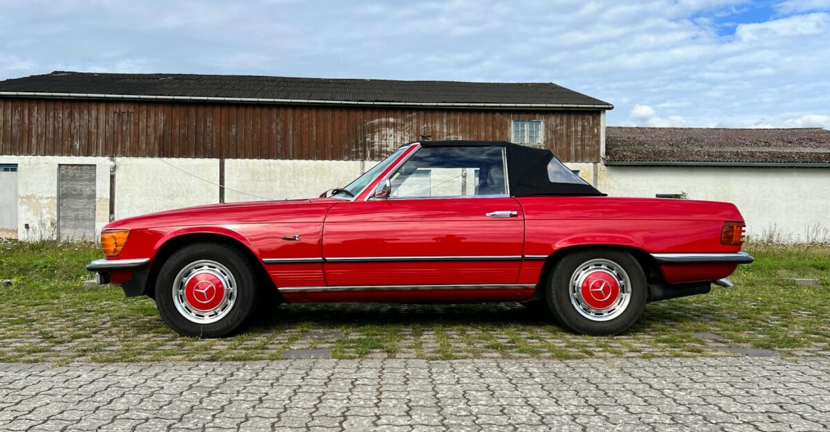 Classic red Mercedes convertible parked on cobblestone by rural wooden barn