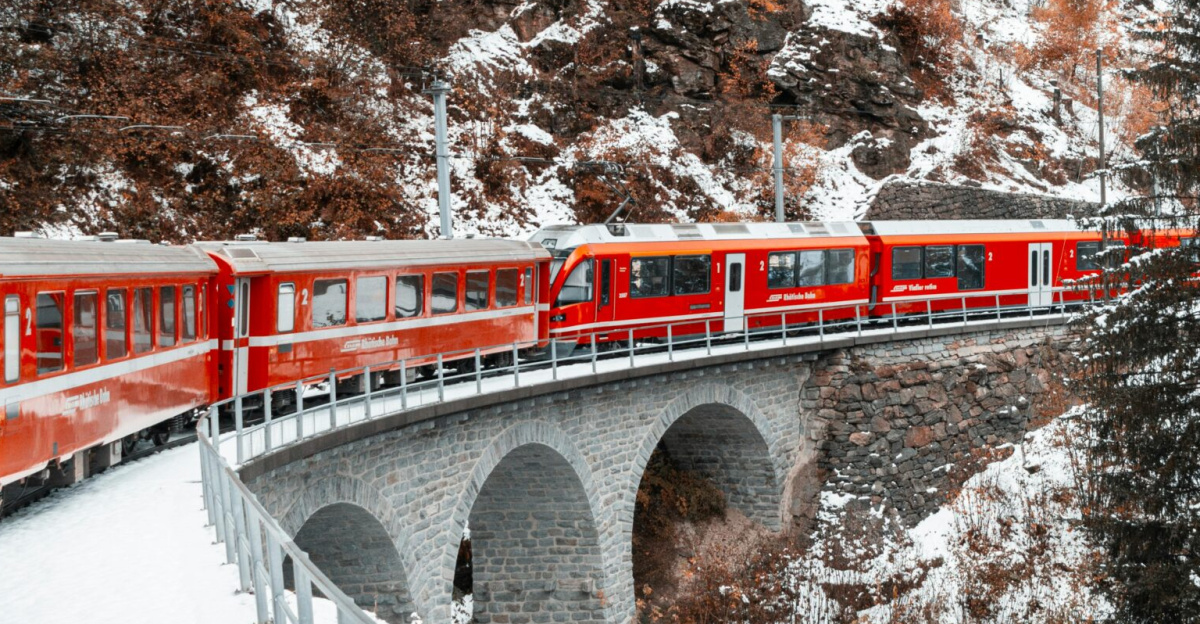 A vibrant red train crosses a snowy coniferous landscape on a scenic mountain bridge in winter.
