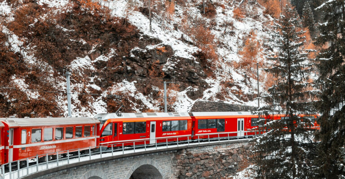 A vibrant red train crosses a snowy coniferous landscape on a scenic mountain bridge in winter.