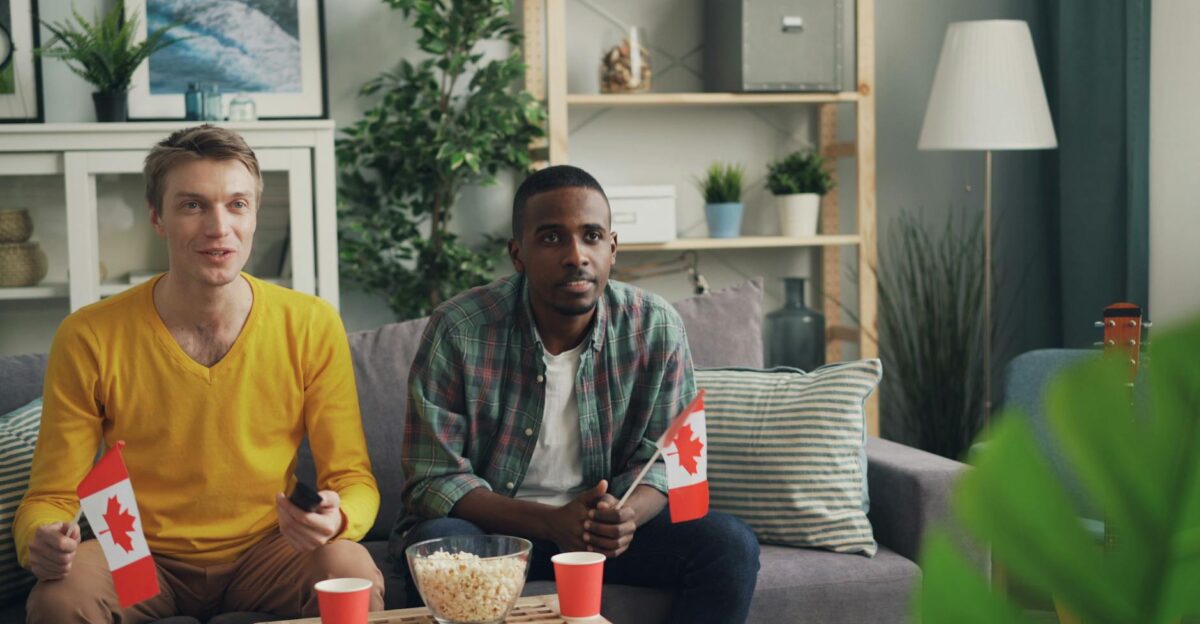 Two friends watching sports on TV with Canadian flags and snacks in a cozy living room