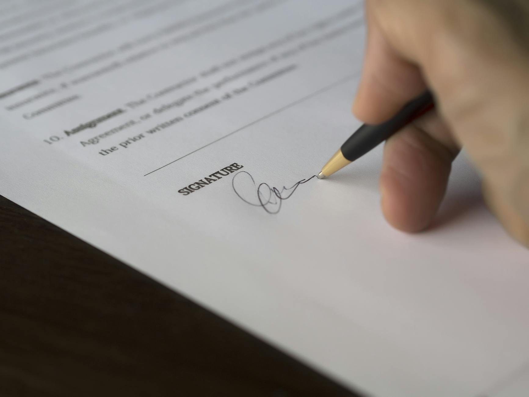 A hand signs a formal contract with a pen on a wooden desk