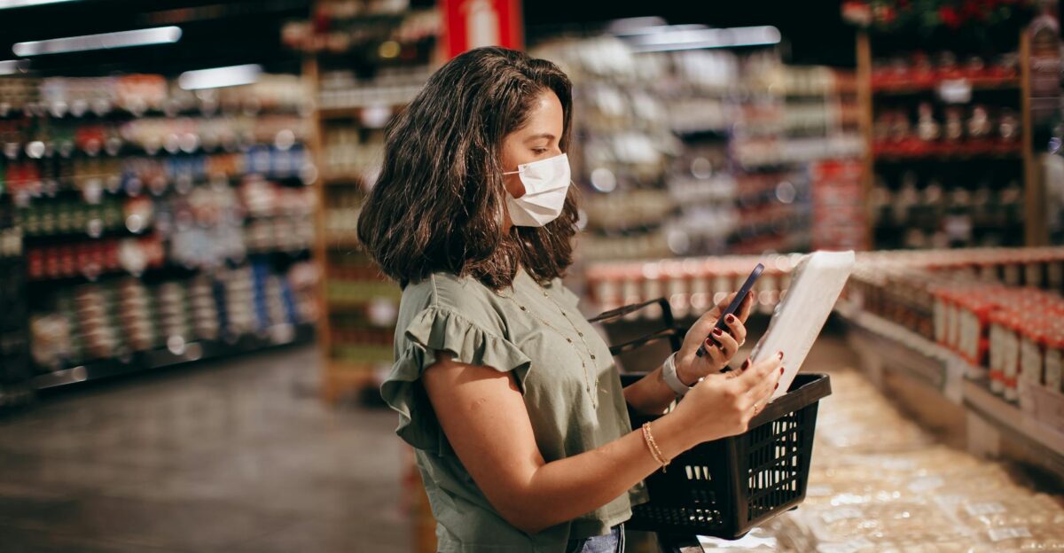 A woman shopping in a supermarket wearing a face mask and using her phone to check products