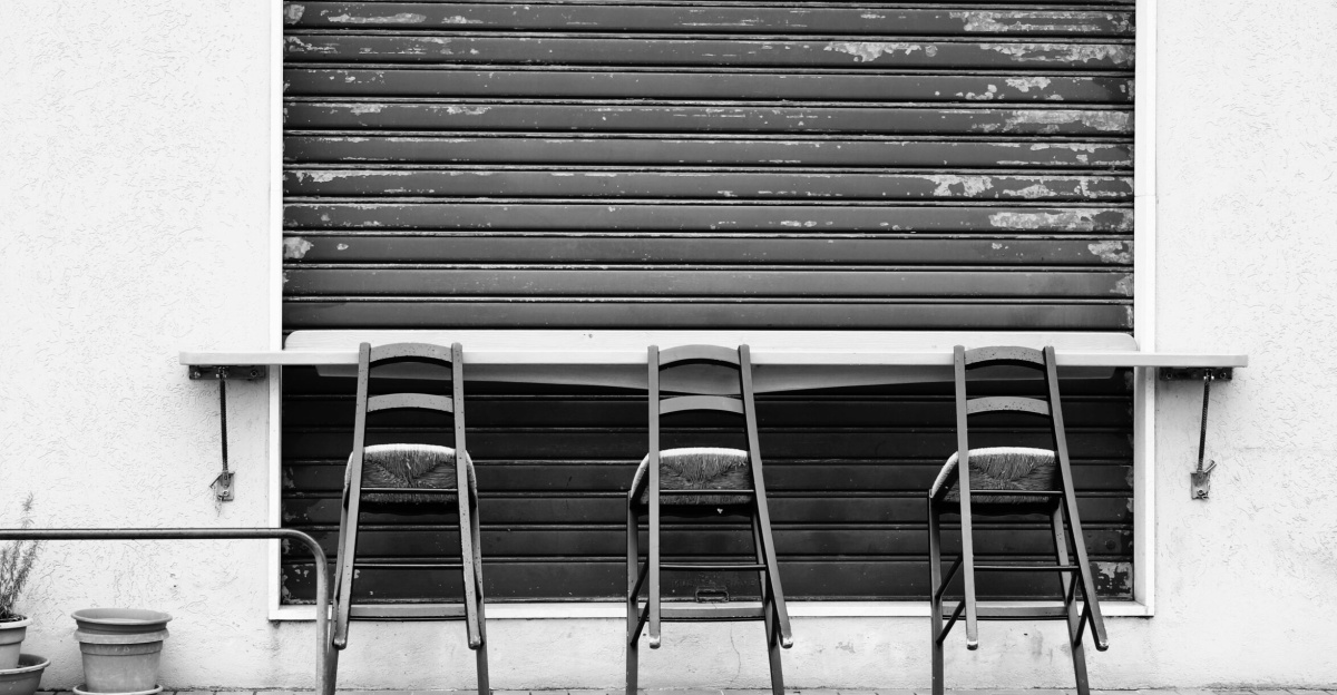 A black and white image of an outdoor cafe with three chairs in front of a closed shutter.