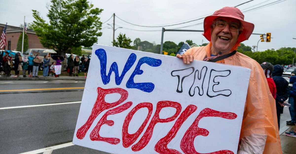A smiling protestor holds a We The People sign during a rally in South Kingstown Rhode Island