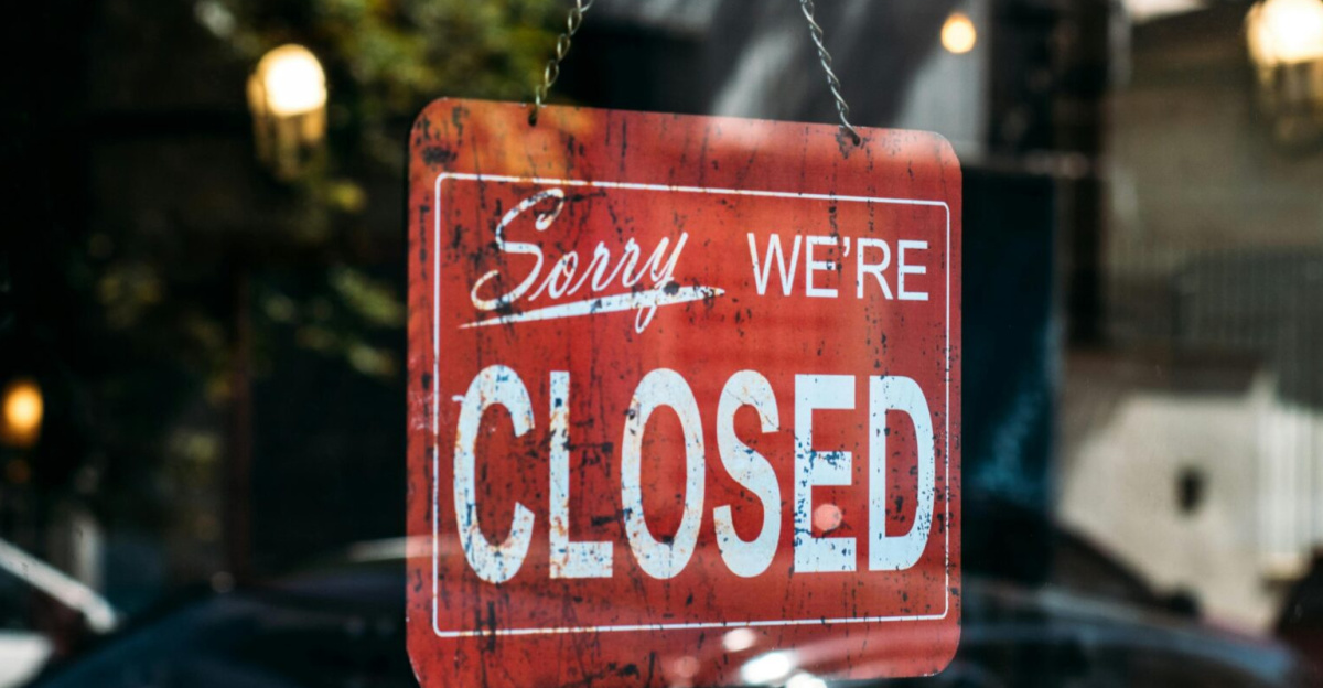 A red closed sign hangs on a glass door, reflecting cars and trees outside.