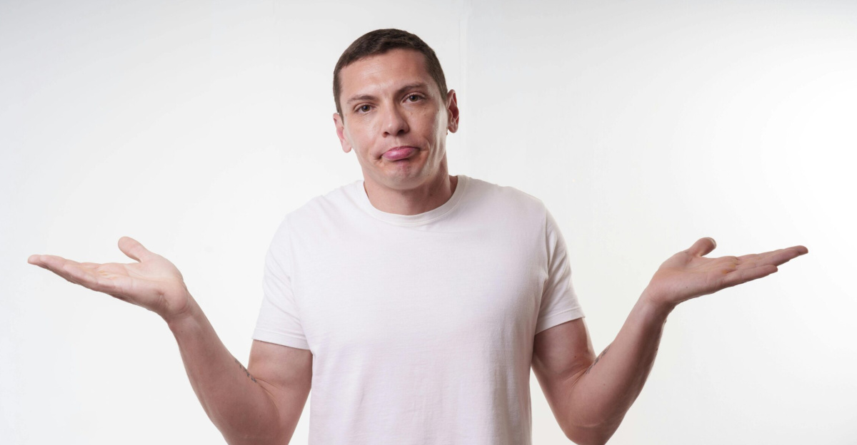 Adult man in white t-shirt shrugs in studio, expressing confusion against a plain background.