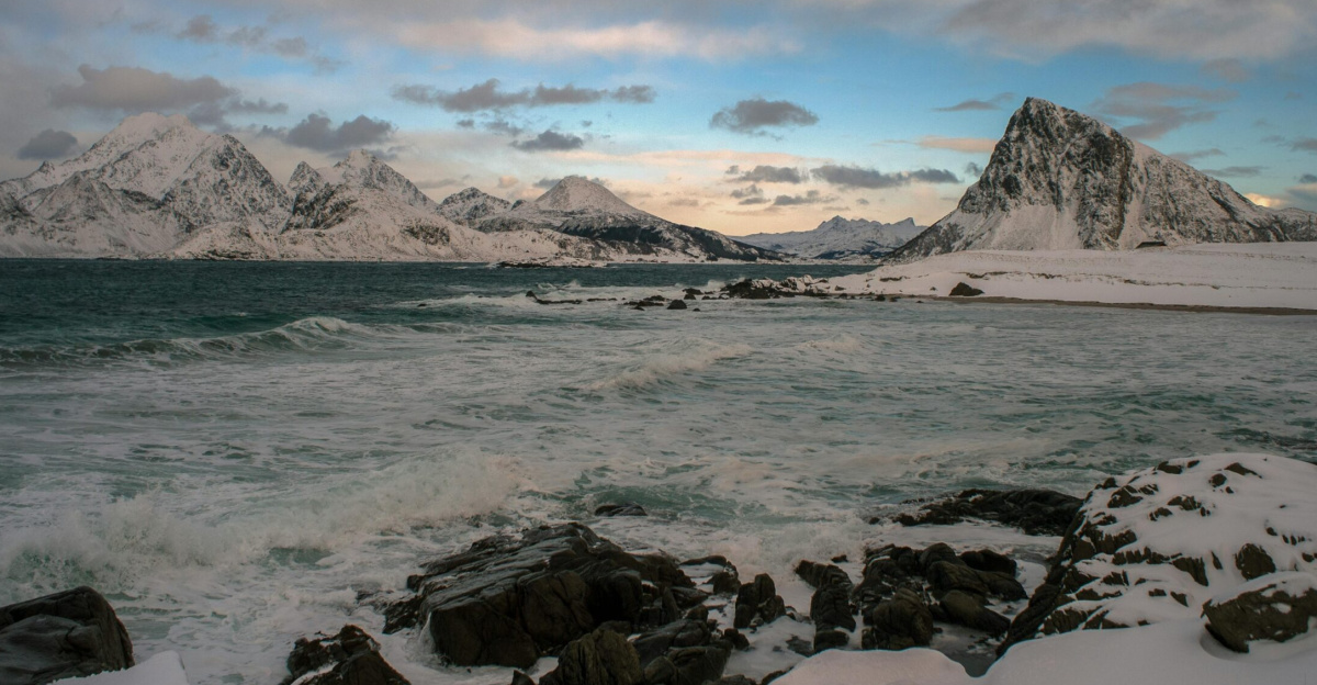 Stunning view of the Lofoten Islands fjord with snow-covered mountains, rocky shore, and cloudy sky.