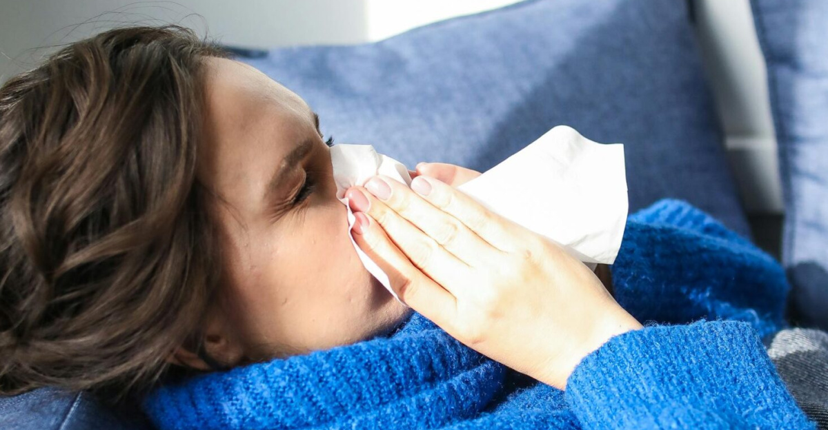 A woman in a blue sweater uses a tissue, lying indoors, suggesting illness or flu symptoms.