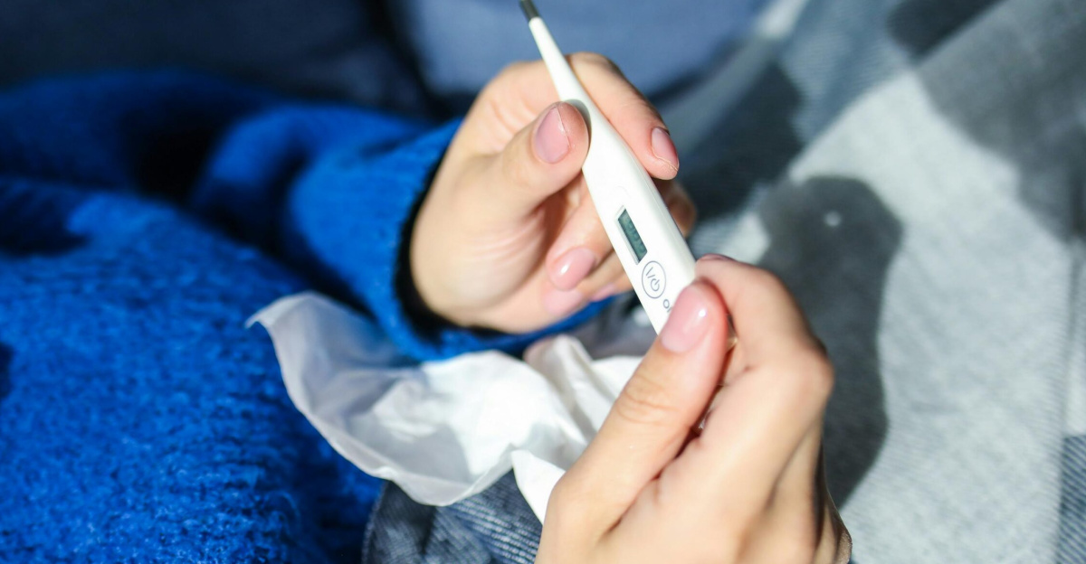 A woman checks a digital thermometer while lying indoors, showing signs of illness.