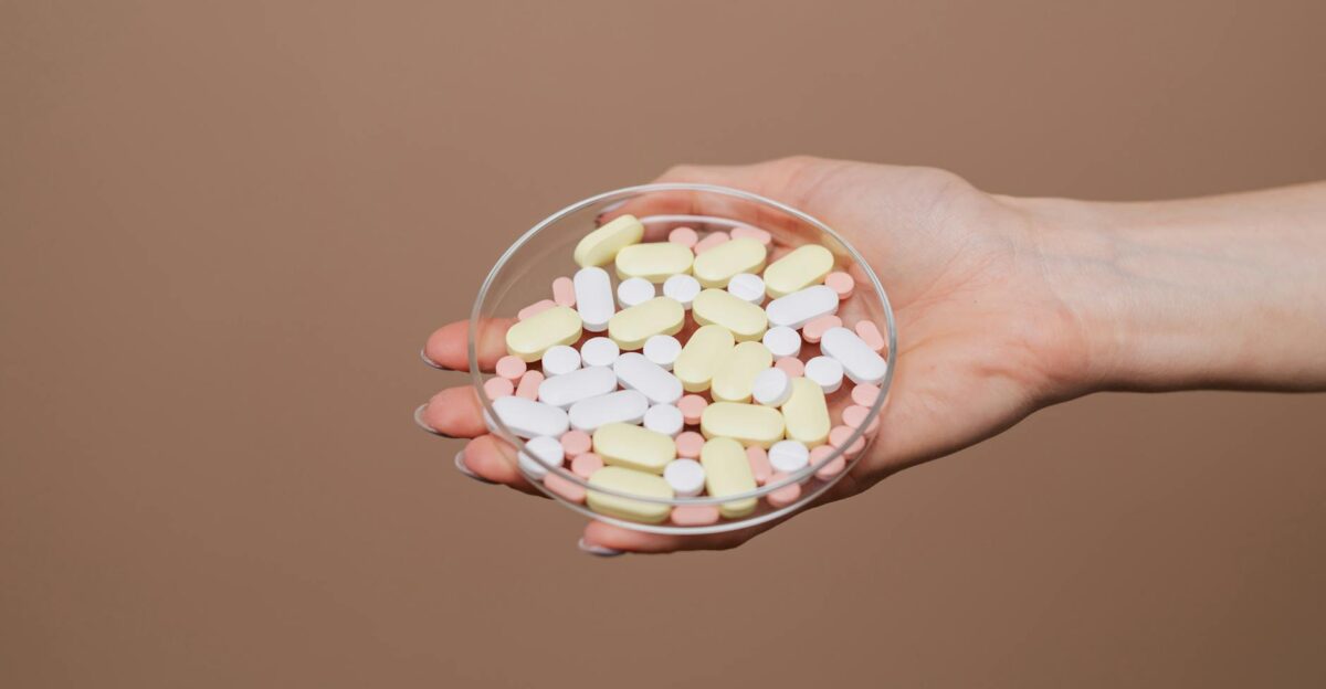 Close-up of a hand holding a petri dish filled with various tablets and capsules on a neutral background