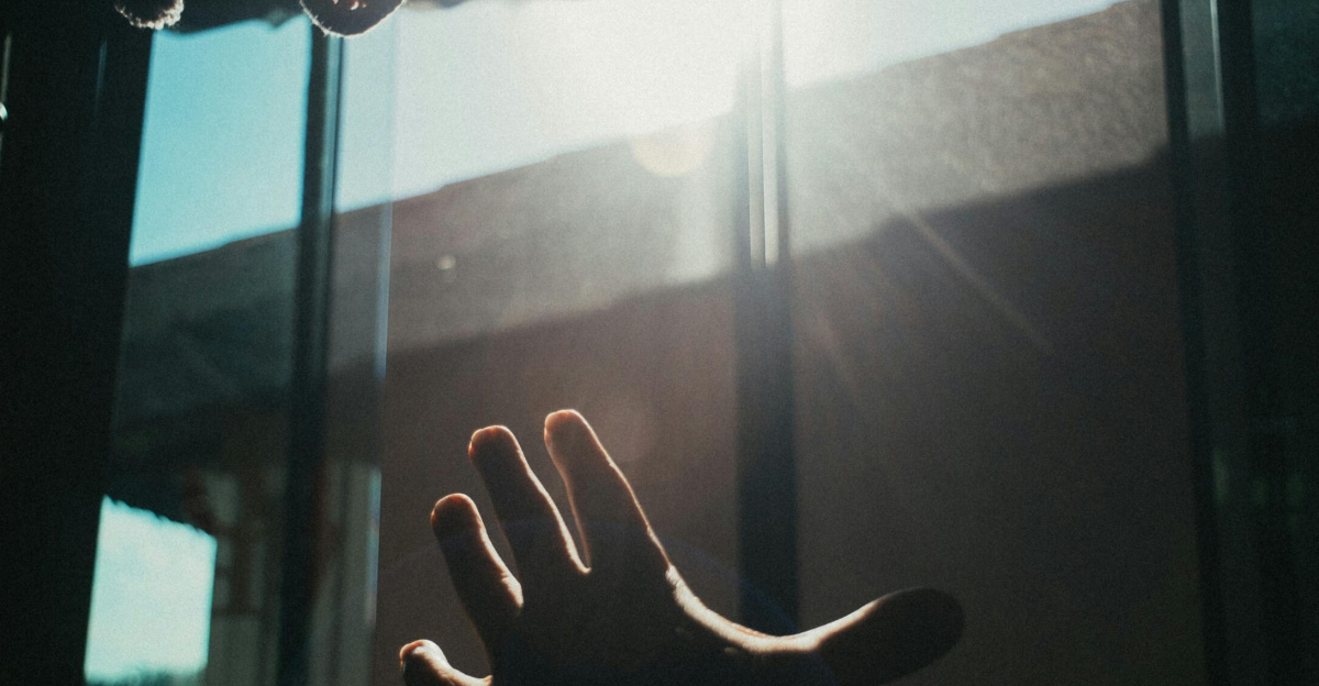 A hand reaches towards the bright sunlight streaming through a window indoors.