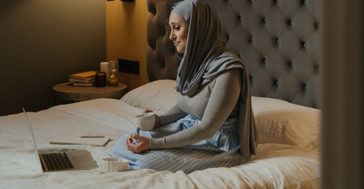 A woman in a hijab sits with her laptop in a warm stylish bedroom drinking coffee and browsing online