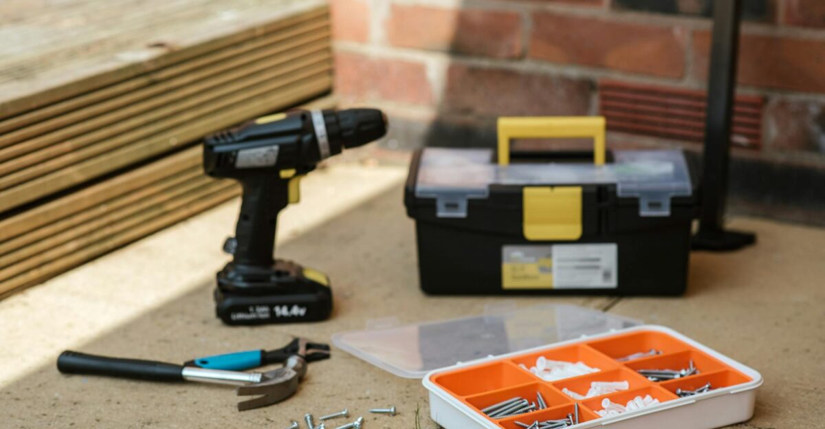 Electric screwdriver on floor near containers with instruments placed near spanner with hammer against brick wall and wooden barrier