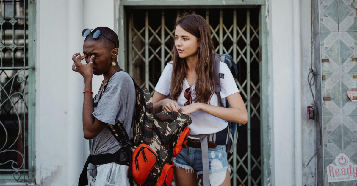 Two travelers with backpacks take a break outside a hotel, showing expressions of fatigue and discussion.