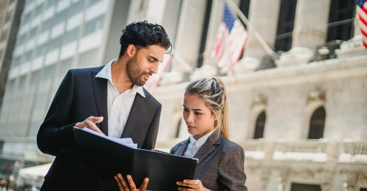 Two professionals in suits discussing documents outside a corporate building in daylight