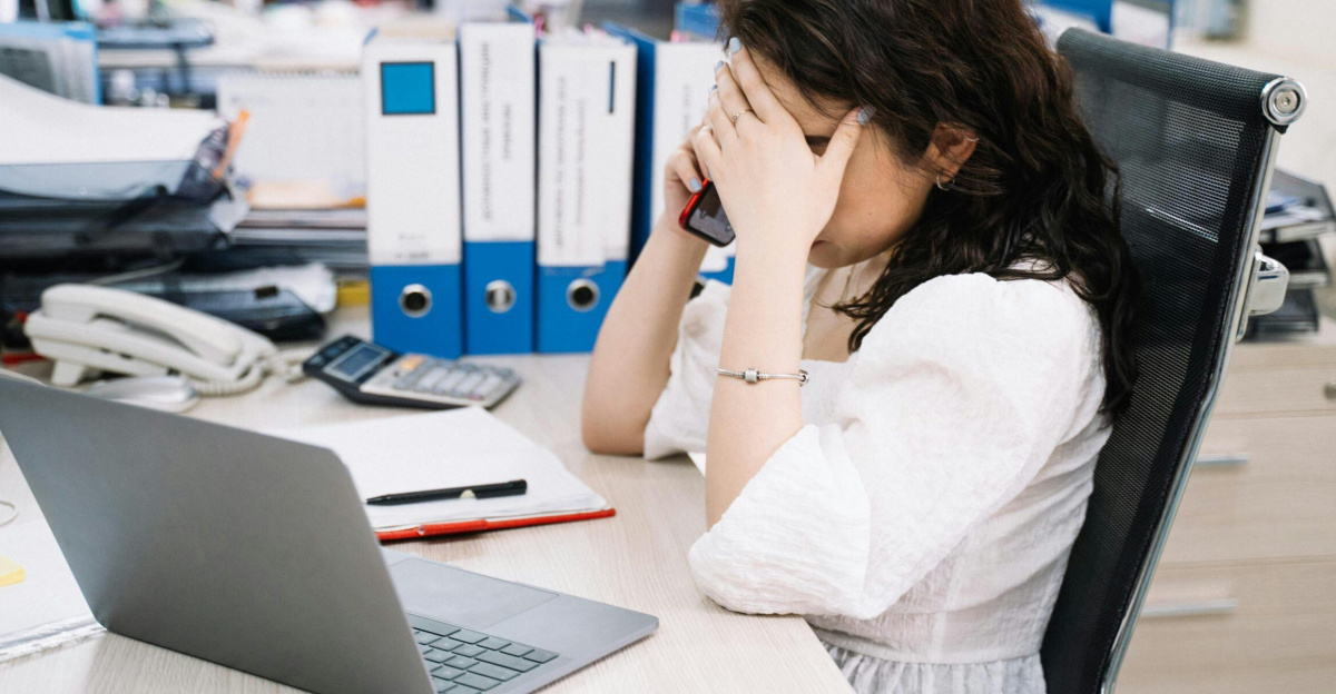 A woman in an office sitting at a desk, visibly stressed, using a phone. Workplace stress concept.