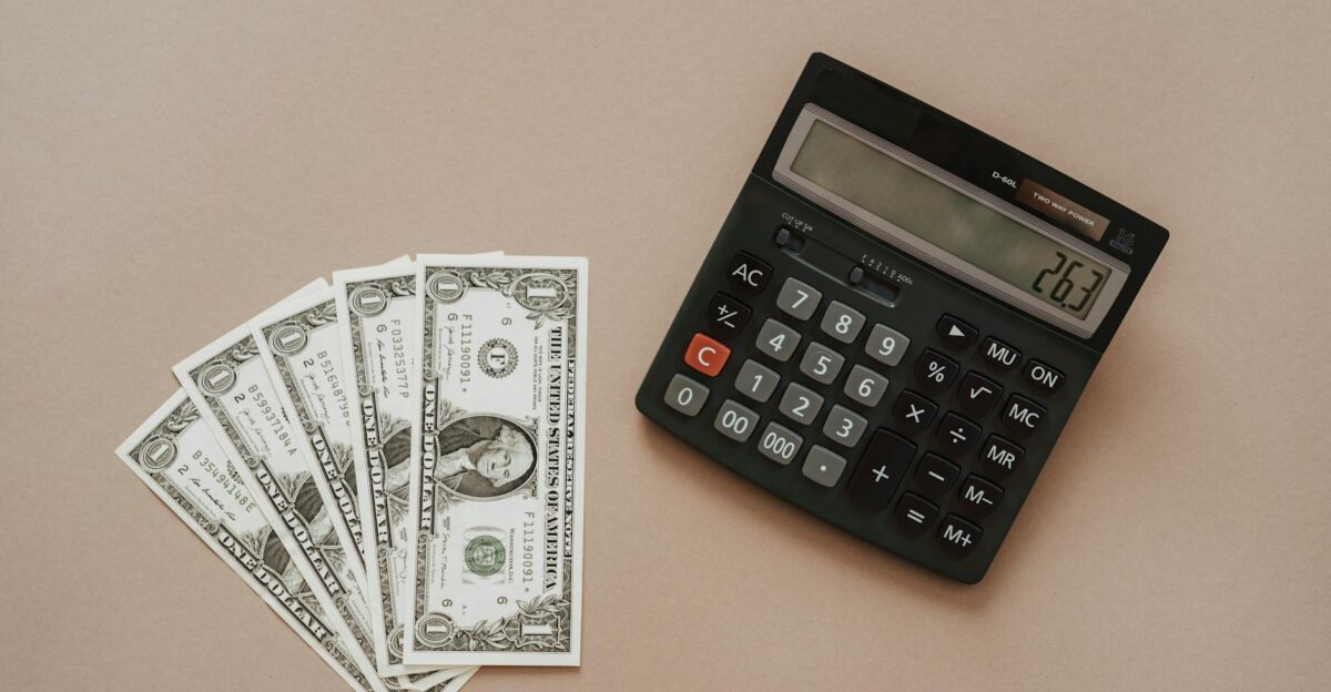 A calculator displaying 26 3 beside four one-dollar bills on a beige background