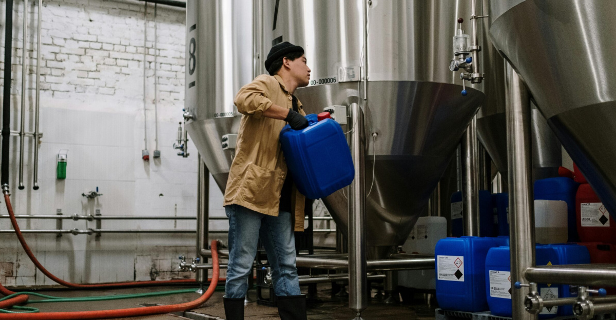 Man handling blue container in a brewery with stainless steel tanks.