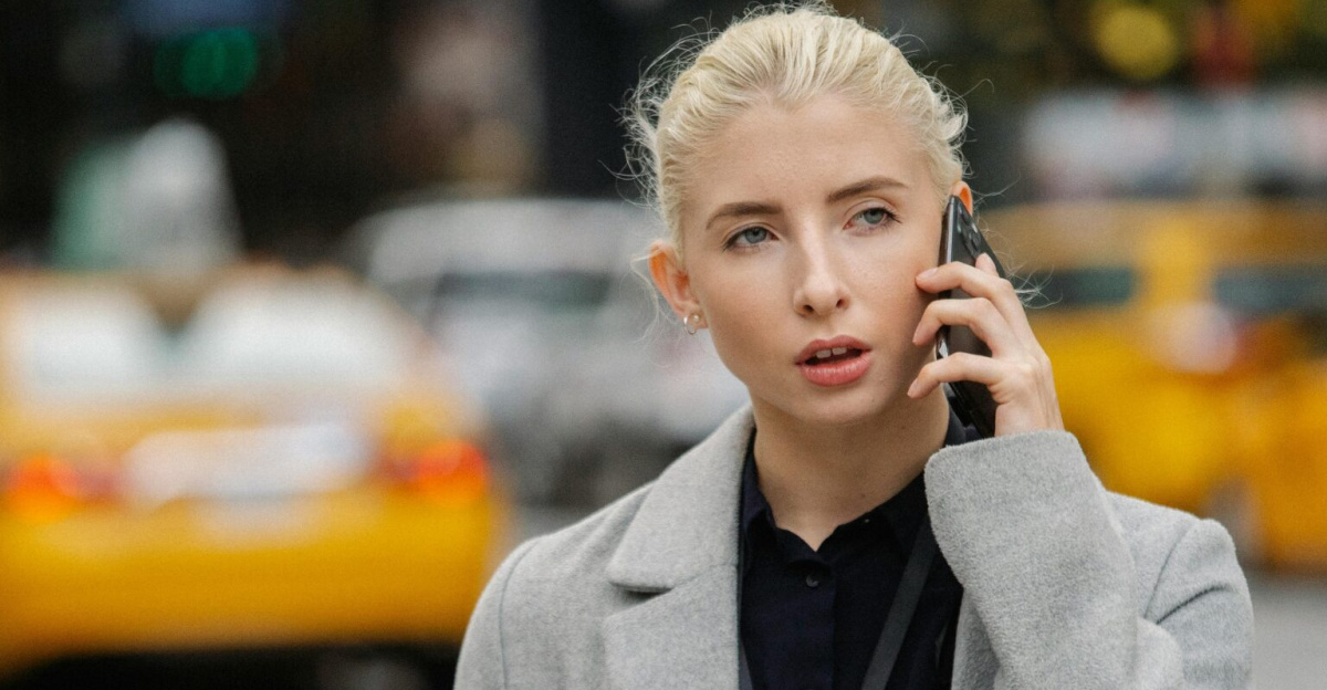 Focused young businesswoman in gray coat standing on busy street with folders and having conversation on mobile phone