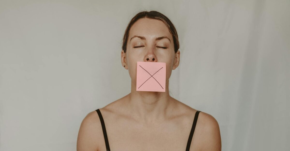 Young slender woman with closed eyes and mouth covered with sticky note showing cross on white background