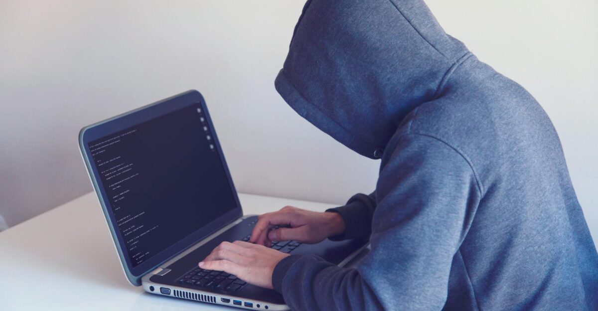 Side view of unrecognizable hacker in hoodie sitting at white table and working remotely on netbook in light room near wall
