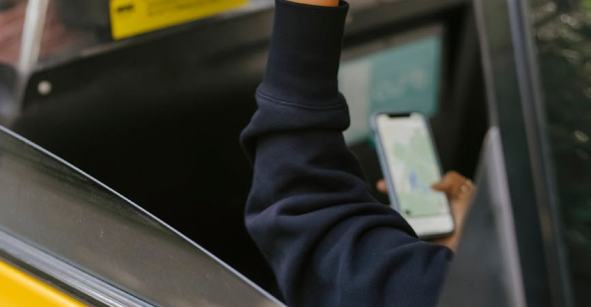 Unrecognizable passenger sitting on backseat and browsing smartphone while holding handle inside of yellow cab with lowered window during ride