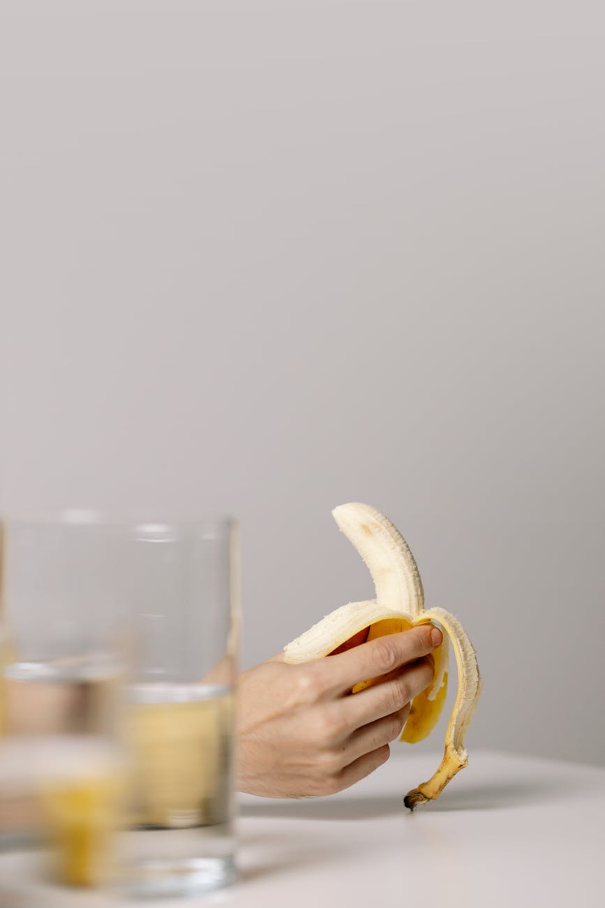 A minimalist photo of a hand holding a peeled banana next to a glass of water on a white surface
