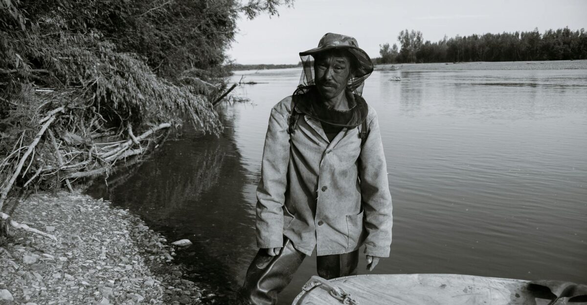 Black and white of calm mature ethnic fisherman in anti mosquito head net hat standing in shallow water of lake near wooden boat and looking at camera